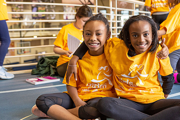Two smiling elementary-aged Girls on the Run participants sitting legs crossed, with an arm around each others shoulders. 